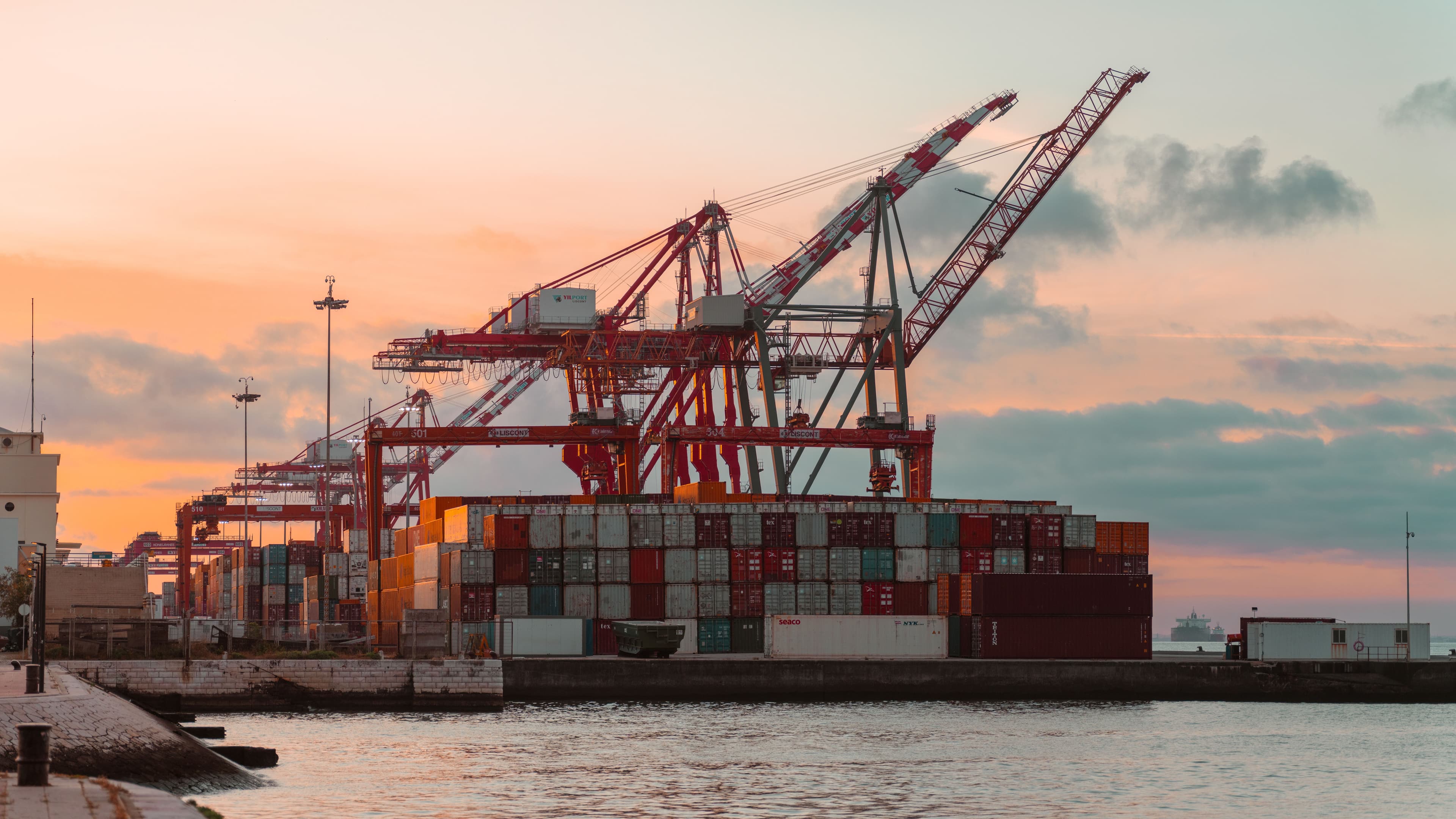 Stacked shipping containers and orange yard equipment at a port terminal.